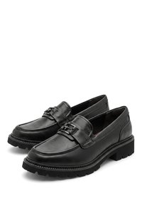 Paire de mocassins en cuir noir avec semelles en caoutchouc épaisses et détail de boucle métallique sur le dessus, présentés sur fond blanc.