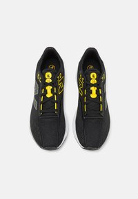 Zapatillas deportivas negras con parte superior de malla, acentos de cordones amarillos y logotipo gris reflectante. Cuentan con una suela acolchada y un sistema de cordones estándar.