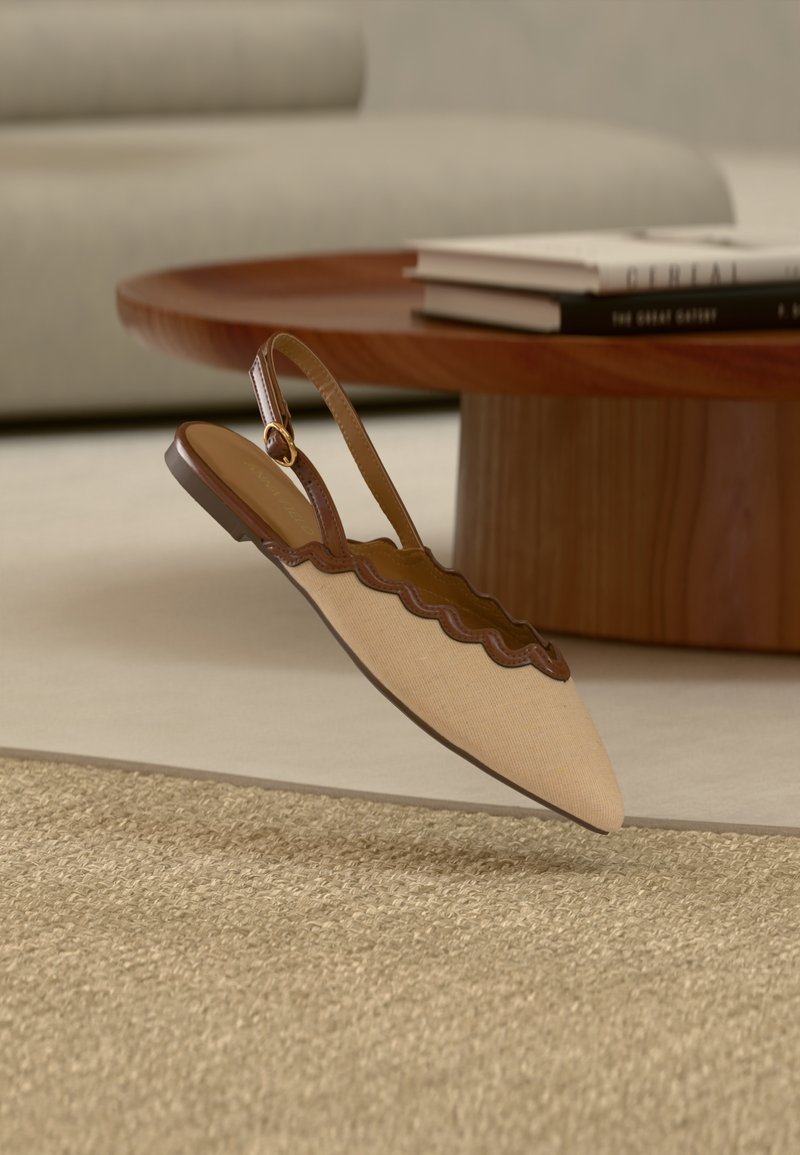 Μπεζ επίπεδη μυτερή γόβα slingback με καφέ ραβδωτό περίγραμμα και λουράκι με αγκράφα, αιωρούμενη πάνω από μπεζ υφασμάτινο χαλί κοντά σε ξύλινο τραπέζι.