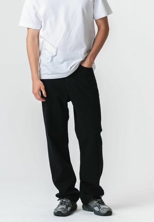 Personne portant une chemise blanche à manches courtes, un pantalon noir et des baskets grises, debout avec une main dans la poche devant un fond uni.