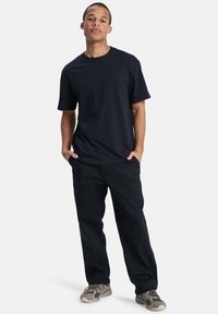 Jeune homme debout avec les mains dans les poches portant un t-shirt noir, un pantalon noir et des baskets grises sur un fond blanc.