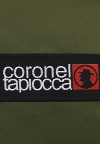 Tela texturizada de color verde oliva con una etiqueta negra que presenta el texto blanco "coronel tapiocca" y un acento rojo con un diseño de silueta.