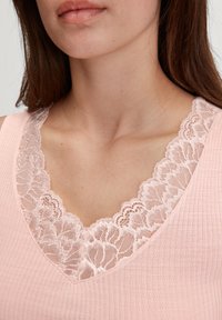 Roze mouwloos topje met een V-halslijn afgewerkt met kant, gemaakt van een gestructureerde stof met een ribbelpatroon, wat de details van de halslijn accentueert.