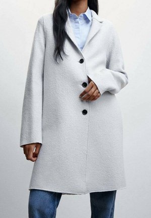 Femme portant un manteau en laine gris clair avec trois boutons noirs sur une chemise bleue et un jean bleu, debout devant un fond uni.