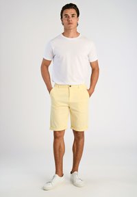 T-shirt en coton blanc à manches courtes associé à un short en coton jaune clair. Le modèle porte des baskets blanches ; le short présente un ourlet roulé.