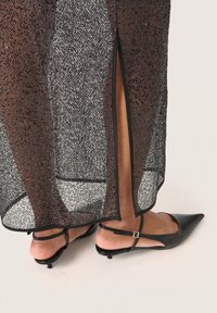 Durchsichtiges schwarzes Kleid mit metallischem Faden und einem seitlichen Schlitz. Kombiniert mit schwarzen spitzen Ballerinas und Knöchelriemen, die die Beinfreigabe betonen.