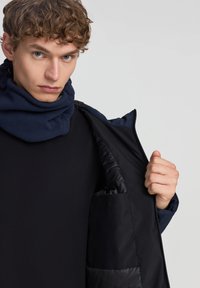 Giacca nera imbottita con sciarpa in pile blu, dotata di chiusura con zip e tasca laterale. Texture liscia con materiali a contrasto. Vestibilità casual.