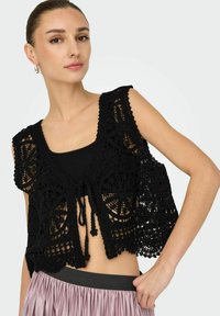 Gilet all'uncinetto nero con motivi floreali, design aperto frontale e chiusura a cravatta, indossato sopra una maglia nera aderente e sovrapposto a una gonna rosa.