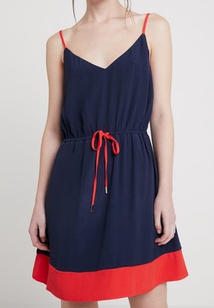 Námořnicky modré šaty bez rukávů s červenými tenkými ramínky, červeným páskem v pase a červeným lemem, které nosí stojící osoba na jednoduchém pozadí.