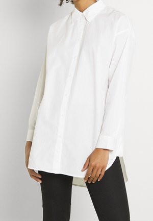 Chemise blanche à bouton avec un col, des manches longues et un ourlet arrière légèrement plus long. Fabriquée dans un tissu léger avec une texture lisse.