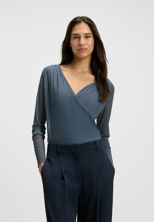 Blue wrap top with long, semi-sheer sleeves, textured fabric, and deep V-neckline, paired with navy striped high-waisted trousers.