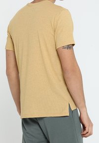 Hombre con una camisa amarilla de manga corta con cortes laterales y pantalones grises, visto de espaldas, mostrando un tatuaje parcial en el brazo.