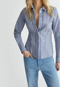 Randig skjorta i blått och vitt, med ett mönstrat knäppband och manschetter, långärmad och figursydd. Bärs med blå jeans.