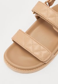 Beige sandaal met twee gequiltte banden, een glad voetbed en een ronde neus. Gemaakt van synthetisch materiaal met minimale stiksels.