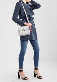 Woman wearing navy trench coat, skinny blue jeans, black high heel sandals, and carrying a white floral crossbody bag with a tassel.