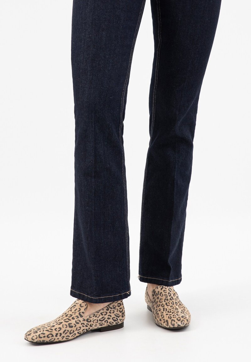 Gambe che indossano jeans in denim blu scuro e scarpe slip-on beige con stampa leopardata, in piedi su una superficie bianca.