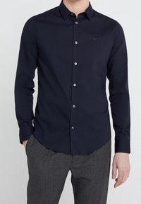 Mans långärmad skjorta i mörk marinblå tyg, med krage med knappar, framknappar och en liten broderad logotyp på bröstet.