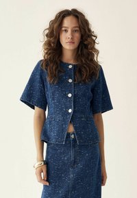 Giacca di denim con maniche corte, texture a pois blu chiaro, bottoni frontali e due tasche. Abbinata a una gonna di jeans coordinata.