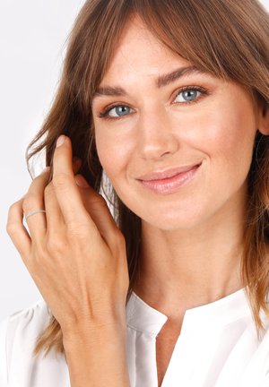 Silver ring on a hand, featuring a simple design and a small accent stone. Model has shoulder-length brown hair and wears a white blouse.