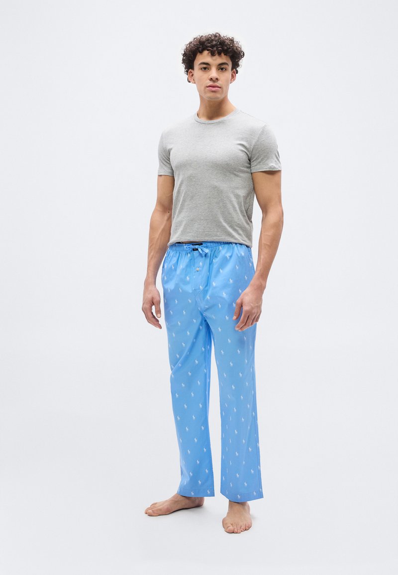 Νέος άνδρας στέκεται ξυπόλυτος φορώντας γκρι μπλουζάκι και ανοιχτόχρωμο μπλε παντελόνι πιτζάμας με μικρό λευκό σχέδιο αλόγου σε λευκό φόντο.