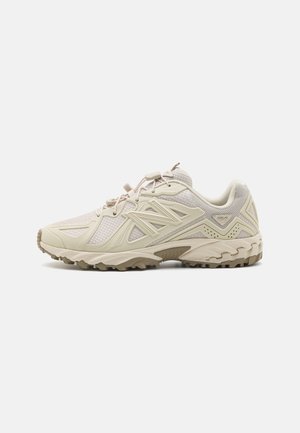 Chaussure de sport beige avec un dessus en maille, des superpositions synthétiques texturées et une semelle en caoutchouc robuste pour l'adhérence. Elle dispose de lacets élastiques et d'un col rembourré.