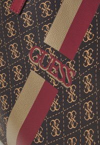 Zbliżenie ciemno-brązowej tkaniny o fakturze z beżowymi i czerwonymi paskami, jasnobeżowym wzorem monogramu oraz czerwonym logo "GUESS" na torbie.