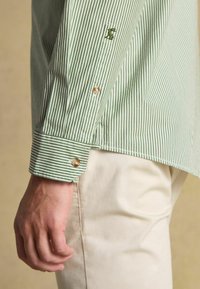 Zeleno-biely pruhovaný košeľový dres s gombíkovými manžetami a vyšitým logom; látka vyzerá hladko s ľahkou textúrou. Krémové nohavice.
