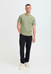 Licht olijfgroen katoenen T-shirt met een ronde hals en een kleine graphic op de linkerborst, gecombineerd met zwarte joggers en sneakers.
