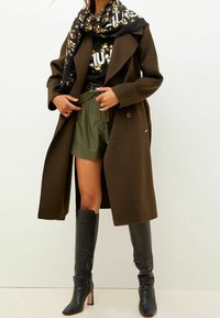 Abrigo de lana marrón con solapas anchas, bufanda negra con estampado, top ajustado negro, pantalones cortos verde oliva y botas de cuero negras hasta la rodilla.