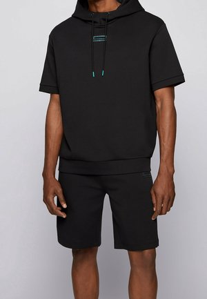 Sort sweatshirt med korte ærmer, hætte og frontlomme; parret med matchende sorte shorts. Har teal-detaljer på snorene.