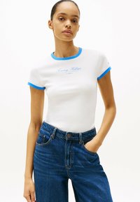 Camiseta blanca de manga corta con cuello y puños de canalé en azul, que presenta el logo de "Tommy Hilfiger" en azul claro. Combinada con unos vaqueros de cintura alta en azul.