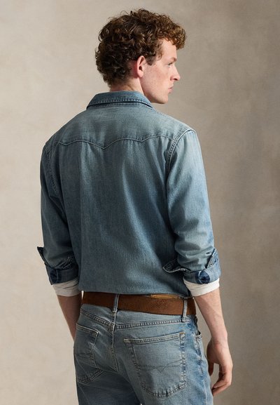Jonge man met krullend haar, gekleed in een lichtblauw denimshirt en spijkerbroek, staand met de rug naar de kijker gekeerd, met opgerolde mouwen en bruine riem.