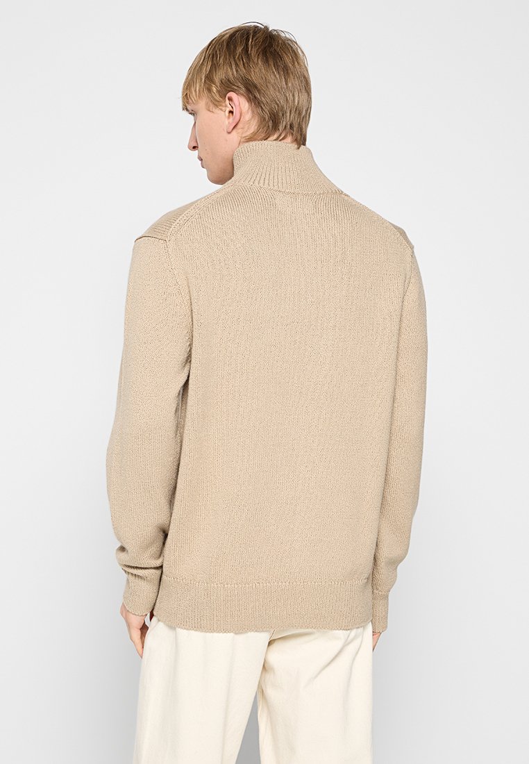 Beige gebreide trui met een hoge kraag, voorzien van een gestructureerd patroon en ribbels bij de mouwen en onderkant. Zichtbare naden geven een extra detail aan het ontwerp.