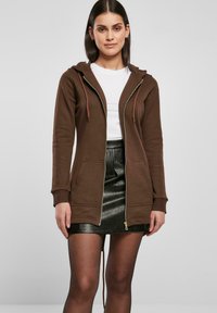 Smeđa zip-up dukserica s prednjim džepovima, s kapuljačom i svijetlim špangama, u kombinaciji s crnom kožnom mini suknjom.