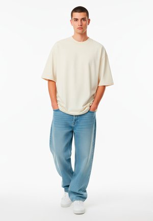 T-shirt oversized color beige chiaro con collo rotondo, abbinata a jeans in denim blu chiaro a vestibilità ampia. Sneakers bianche completano l'outfit.