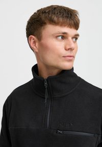 Pullover in pile nero con colletto alto, chiusura frontale a mezza zip e tasca sul petto. Presenta una superficie testurizzata e un marchio discreto nel tessuto.