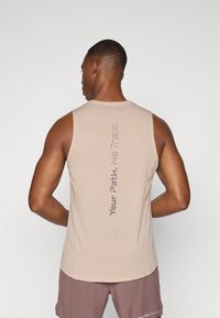 Beige ärmelloses Sporttop aus texturiertem Stoff. Auf der Rückseite befindet sich vertikaler Drucktext. Kombiniert mit braunen Shorts.