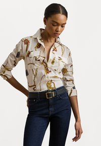 Blusa bianca di cotone con un motivo oro e rosso, design con bottoni e due tasche frontali. Abbinata a jeans in denim blu scuro e una cintura grigia.