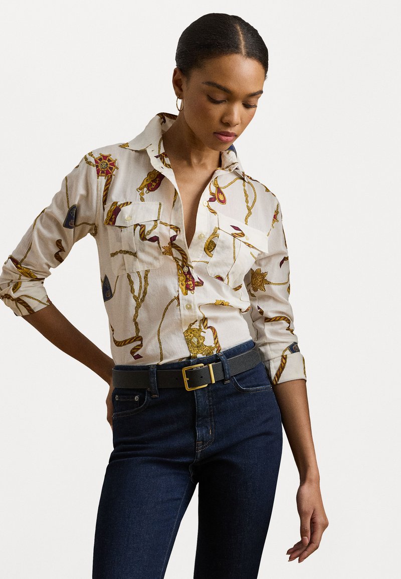 Blusa bianca di cotone con un motivo oro e rosso, design con bottoni e due tasche frontali. Abbinata a jeans in denim blu scuro e una cintura grigia.
