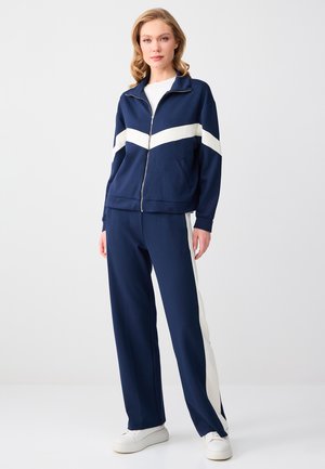 Frau trägt marineblaue Jacke mit Reißverschluss und weißem Chevron-Streifen sowie passende Hose mit weißen Seitenstreifen, steht vor schlichtem Hintergrund.