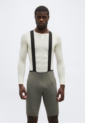 Mužský model v bielej košeli s dlhým rukávom a sivých cyklistických krátkych nohaviciach s čiernymi trakovými popruhmi, stojaci pred svetlosivým pozadím.