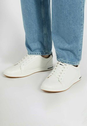 White sneakers made of textured synthetic material, featuring a flat sole, rounded toe, lace-up design, and subtle side accents.