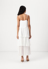 Robe maxi blanche à volants avec des bretelles spaghetti ajustables, fabriquée en tissu léger texturé, comportant trois volants superposés, longueur cheville.