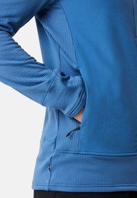 Blauer Pullover mit langen Ärmeln und strukturierten Paneelen, ausgestattet mit einer seitlichen Reißverschlusstasche und einem Flaggenakzent nahe dem Saum. Dehnbarem Stoff für zusätzlichen Komfort.
