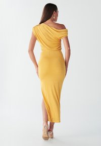 Robe jaune épaules dénudées en tissu lisse, silhouette ajustée, longueur mi-mollet, avec une fente latérale, et chaussures transparentes à talons hauts.