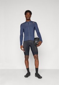 Homme portant un maillot de cyclisme bleu marine à manches longues et un short rembourré noir, tenant un casque de vélo noir sur le côté, se tenant devant un fond uni.