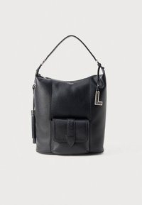 Mala de balde em pele preta com alça superior, bolso com aba frontal, detalhe de tassel e pendente prateado em forma de "L" em fundo simples.