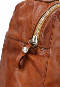 Angolo di uno zaino in pelle marrone con una zip argento prominente e un tirante in pelle. Presenta cuciture visibili e una texture sottile.