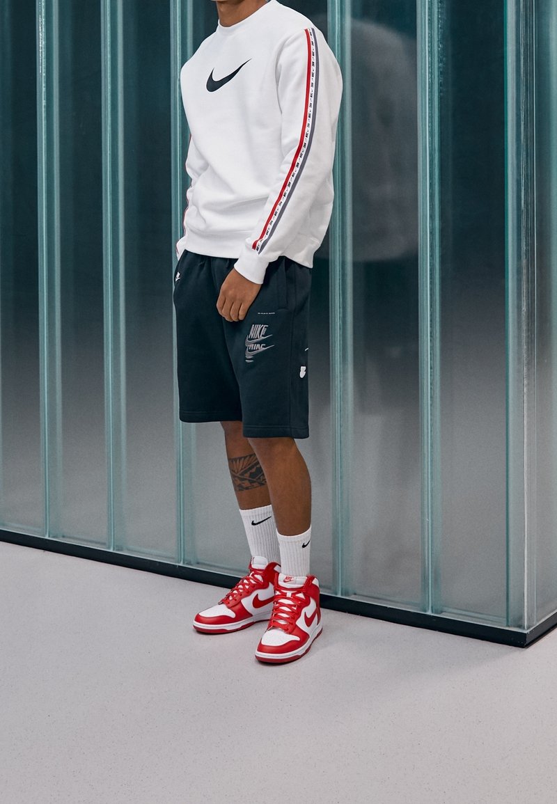 Vit sweatshirt med svart logotyp och röda accenter, mörka shorts med tryckt mönster, röda och vita hög-top sneakers och vita strumpor.