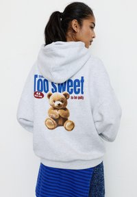 Felpa grigia con cappuccio caratterizzata da una grande grafica di un orsetto. Il testo recita "Troppo dolce per essere colpevole." Tessuto spesso con una texture morbida.